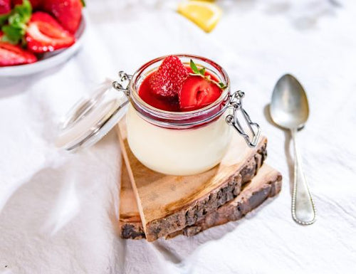 Creamy lemon posset topped with fresh Harry's Berries strawberries in a glass jar, garnished with mint and served with a spoon on a light marble surface.