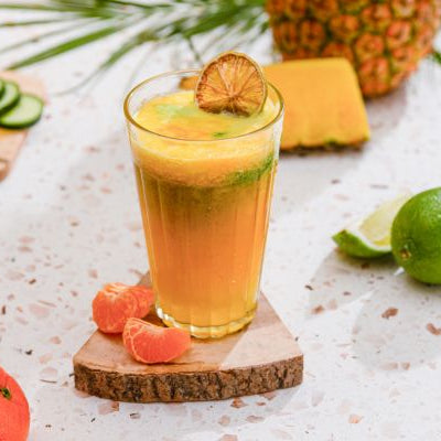 Tall glass of Tropical Glow Juice garnished with a dried orange slice, with pineapple, lime, cucumber slices, and mandarin segments on the table.