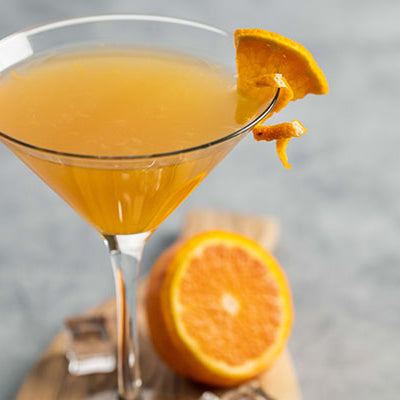 The Bronx – Pixie Tangerine Style cocktail in a martini glass with a gray background and halved tangerine resting against the glass.