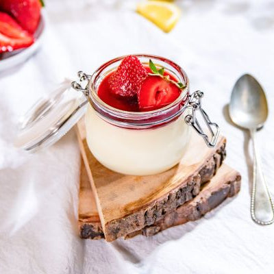 Creamy lemon posset topped with fresh Harry's Berries strawberries in a glass jar, garnished with mint and served with a spoon on a light marble surface.