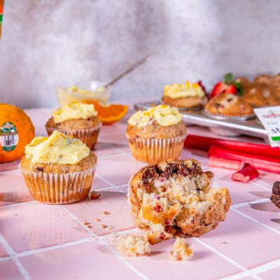 Strawberry rhubarb muffins with tangerine buttercream, one broken open, surrounded by fresh fruit on a pink surface.