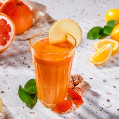 Glass of bright orange turmeric citrus juice garnished with an apple slice, surrounded by grapefruit, lemons, apples, and fresh turmeric root