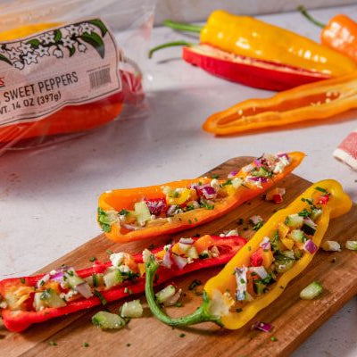 Colorful long sweet peppers halved and filled with a chopped pepper salad, feta cheese, cucumber, red onion, and fresh herbs, served on a wooden board with additional sweet peppers in the background.