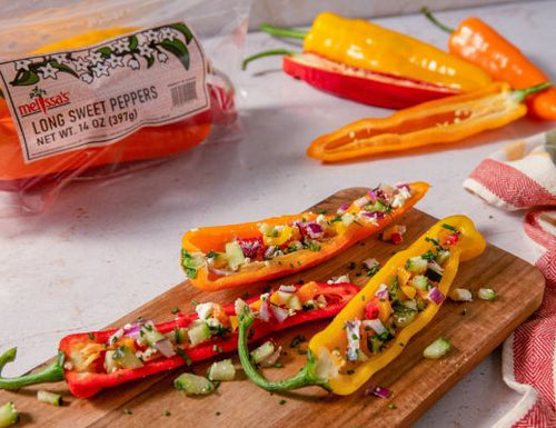 Colorful long sweet peppers halved and filled with a chopped pepper salad, feta cheese, cucumber, red onion, and fresh herbs, served on a wooden board with additional sweet peppers in the background.