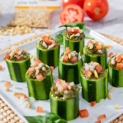 Cucumber cups filled with tropical basil pesto and topped with diced tomatoes and nuts on a white serving plate.