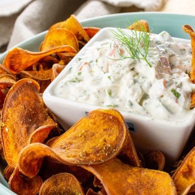 Crispy spiced garnet yam chips served with creamy cucumber herb dip in a white bowl.