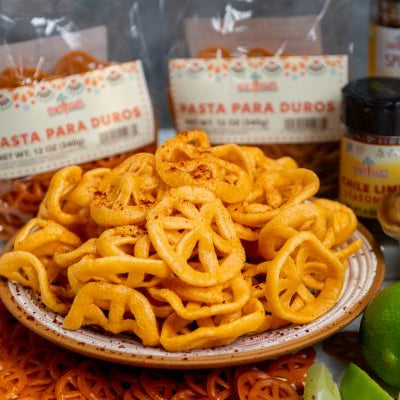 Pasta para Duros Mexican wagon wheel pasta snack piled on a plate, crispy fried duritos seasoned with chili lime, with packaged Pasta para Duros bags and lime wedges in the background