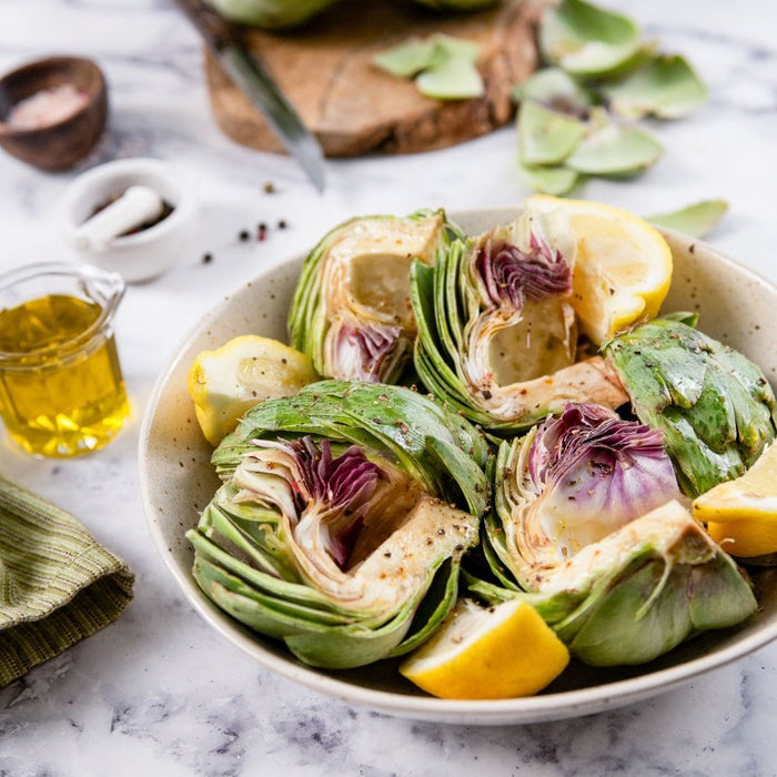Organic Artichokes cut into chunks and seasoned in a bowl with sliced lemons on a white marble surface.