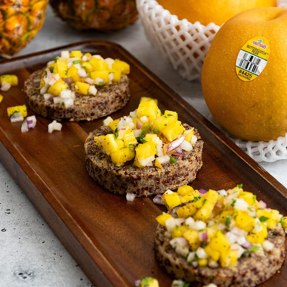 Three small brown quinoa rounds topped with diced yellow mango, white onion, and green herbs, arranged on a dark wooden serving board. Whole pineapples and oranges in white netting sit blurred in the background on a light gray stone surface.