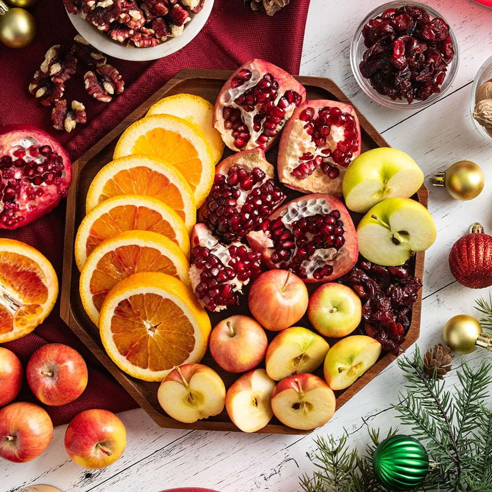 A festive holiday fruit platter arranged on a wooden board with sliced oranges, pomegranates, apples, dried cranberries, and nuts, styled on a white wood surface with Christmas ornaments, evergreen branches, and a red cloth.