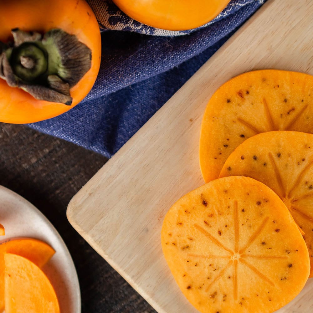 Sliced persimmon on a wooden cutting board and a small plate, with whole persimmon fruits and a blue patterned cloth in the background.