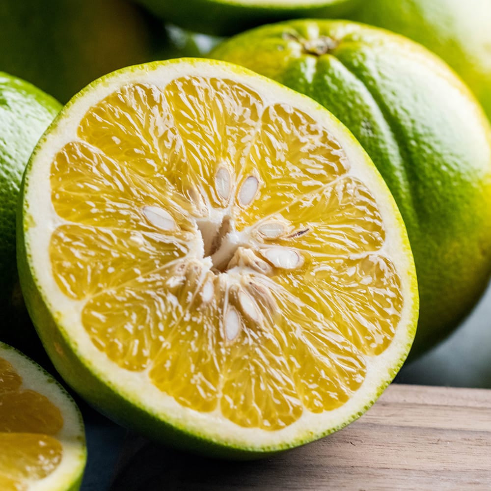 Fresh green citrus fruits, including whole and halved oranges, arranged on a wooden surface with a blurred gray background.