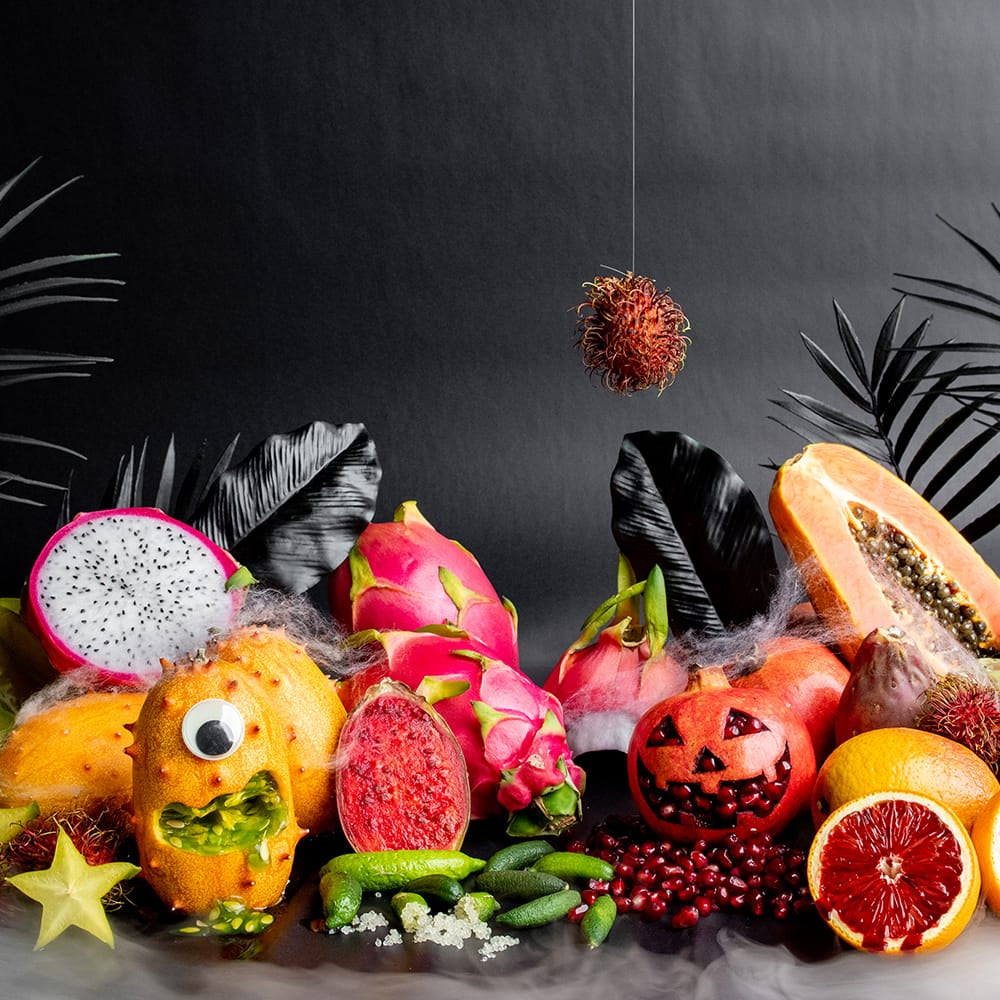 A Halloween-themed fruit arrangement on a dark background, featuring carved fruits like a jack-o'-lantern papaya, dragon fruit, rambutan, and starfruit, along with whole and sliced citrus and passion fruit, with black palm leaves and a hanging red berry.