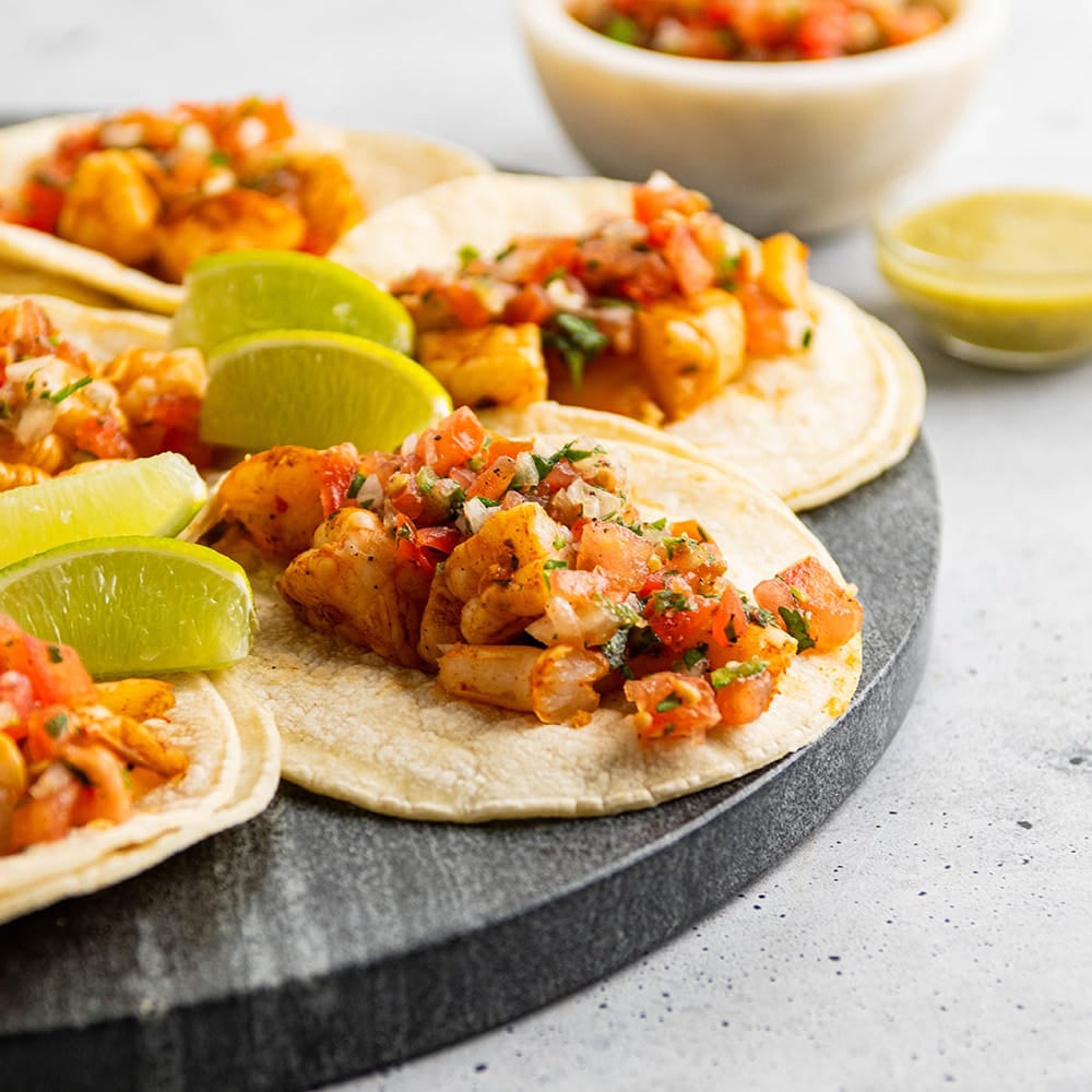 Four shrimp tacos on a dark round serving board, garnished with lime wedges and cilantro, with small bowls of sauce in the background on a light gray surface.
