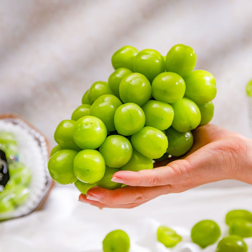 A hand holding a cluster of bright green grapes, with a packaged bag of grapes and a small glass bowl of grapes on a light draped background.
