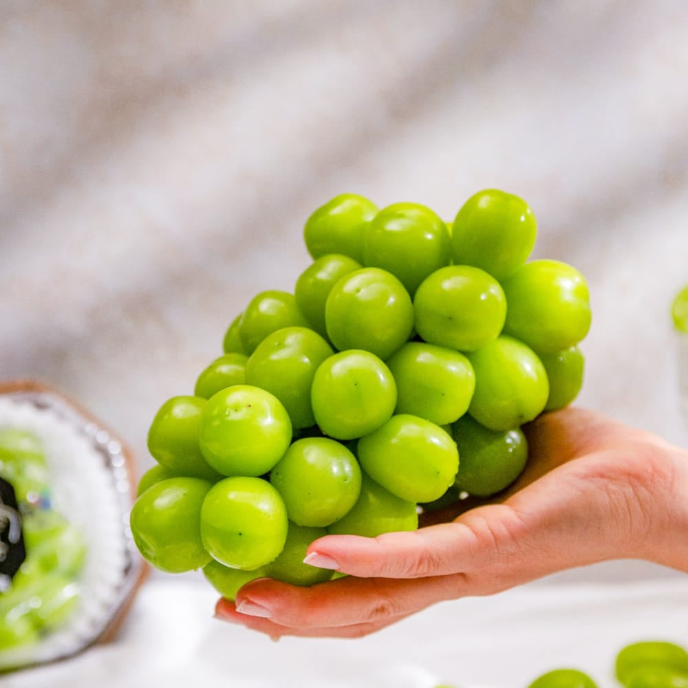 A hand holding a cluster of bright green shine muscat grapes, with a packaged bunch of grapes and a small glass bowl of grapes in a softly lit kitchen-like setting.