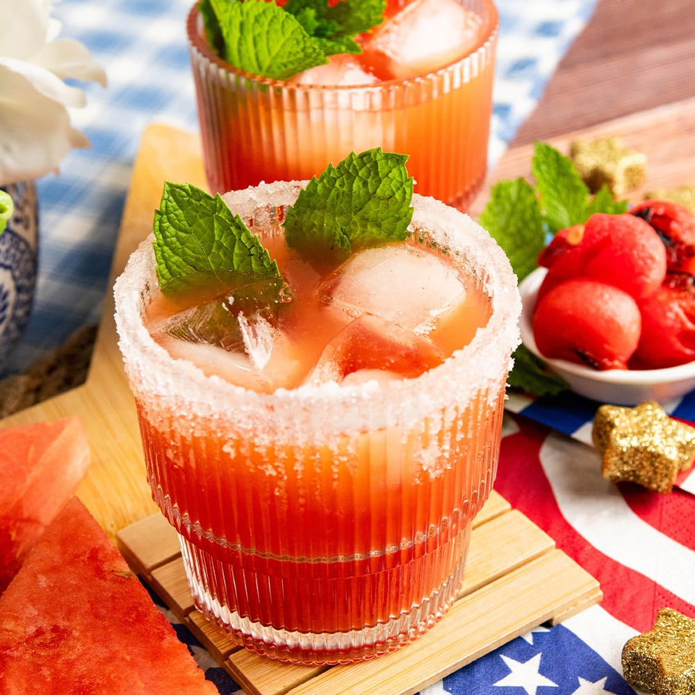 Two sugar-rimmed glasses filled with a pink-orange cocktail, garnished with mint leaves, styled on a wooden surface with watermelon slices, strawberries, a lime, and patriotic napkins.