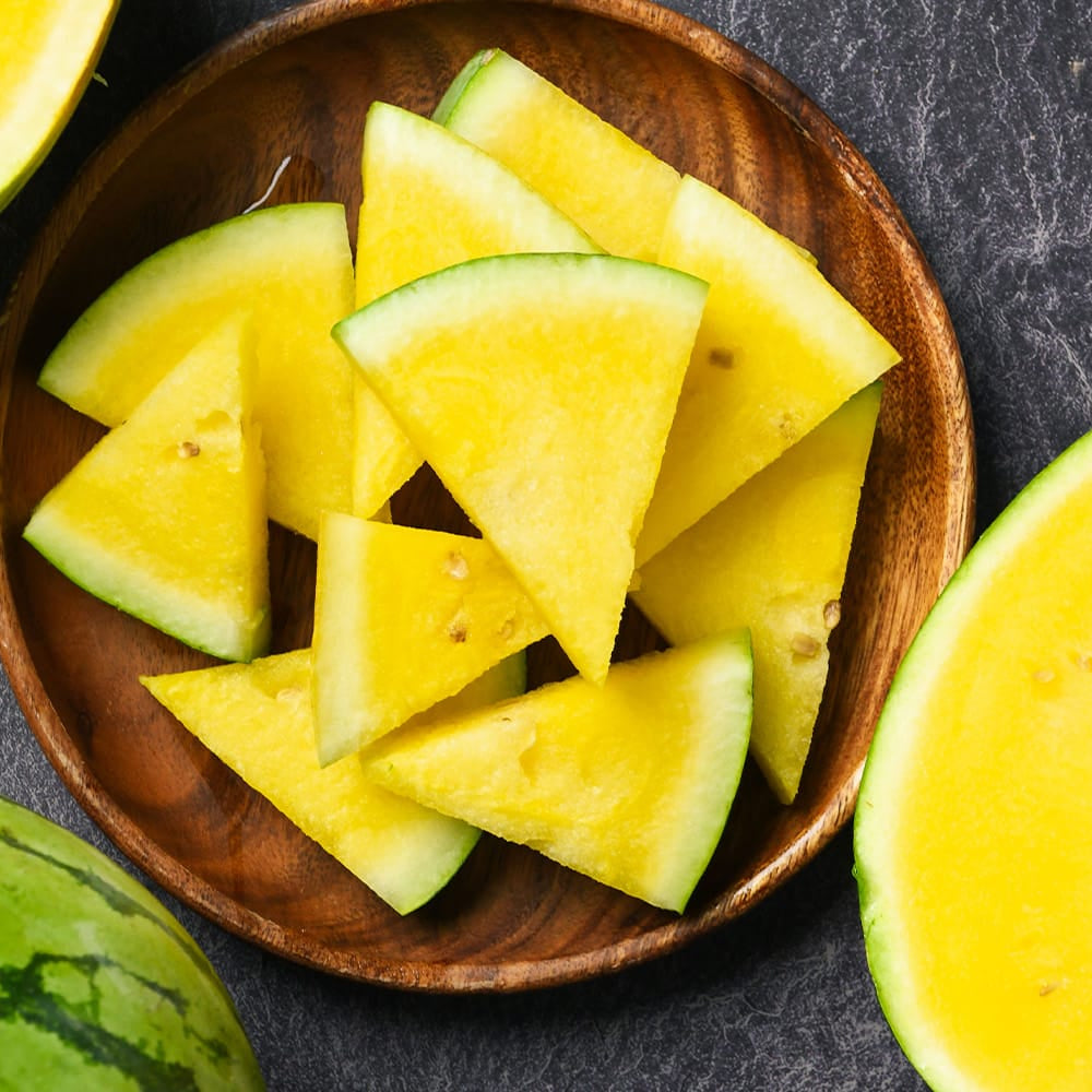 Yellow watermelon slices on a wooden plate with whole yellow watermelons on a dark surface