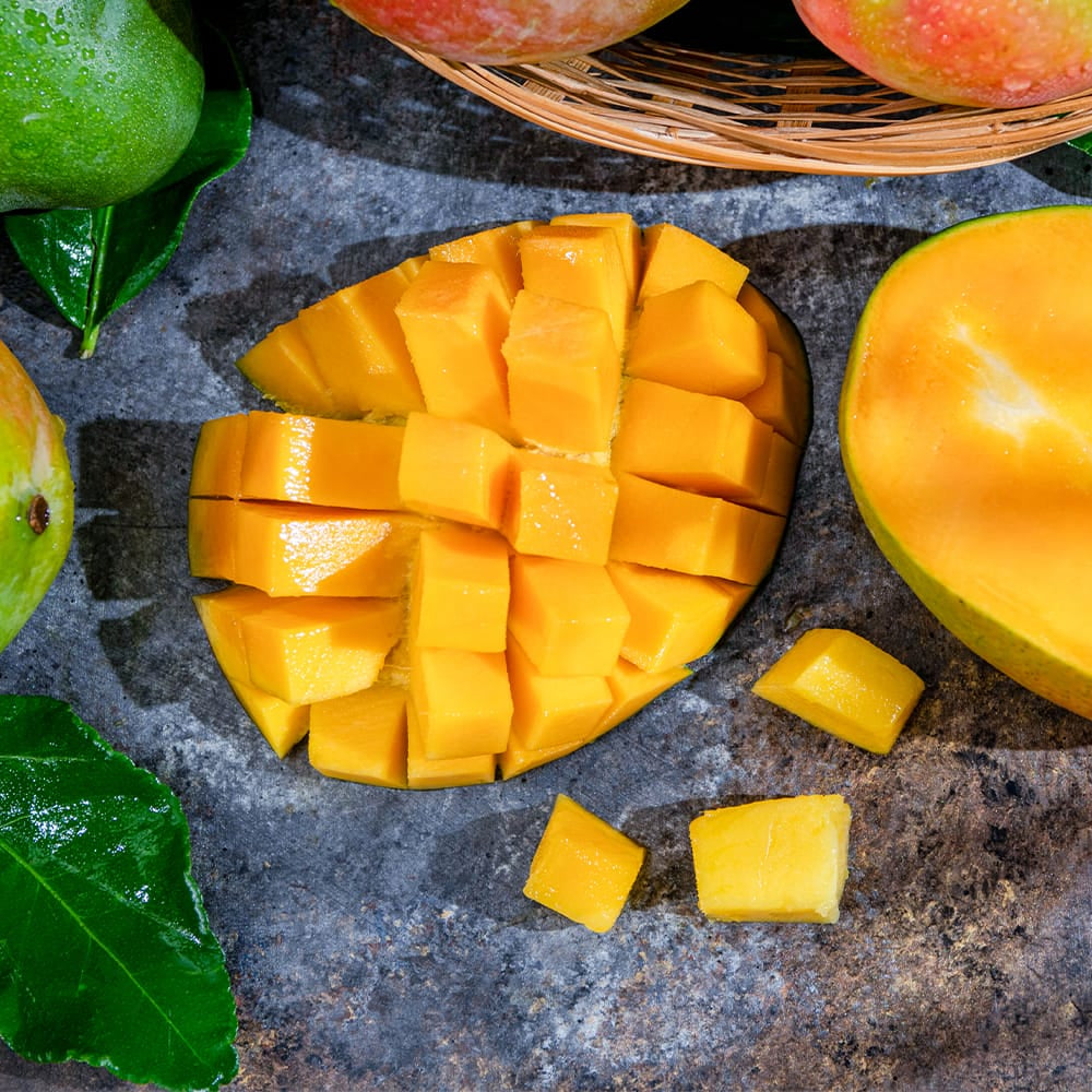 Fresh mangoes arranged on a gray stone surface, including whole green mangoes, a cut mango half showing bright yellow-orange flesh, and a sliced mango cube arrangement in the center, with green leaves scattered around.