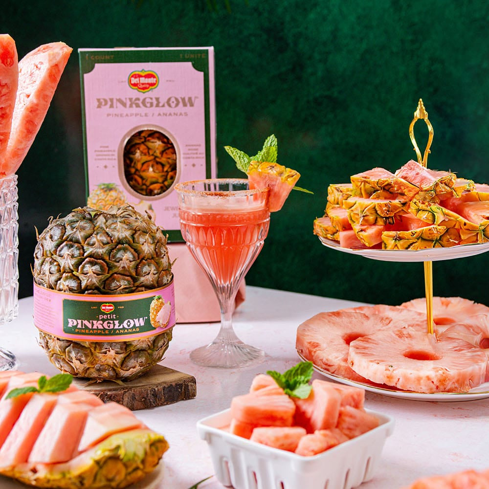 A styled fruit and dessert display featuring Pinkglow branded packaged pink fruit slices, a whole pineapple wrapped in a Pinkglow label, a glass of pink drink, and multiple fruit platters and cups arranged on a white table against a dark green backdrop.