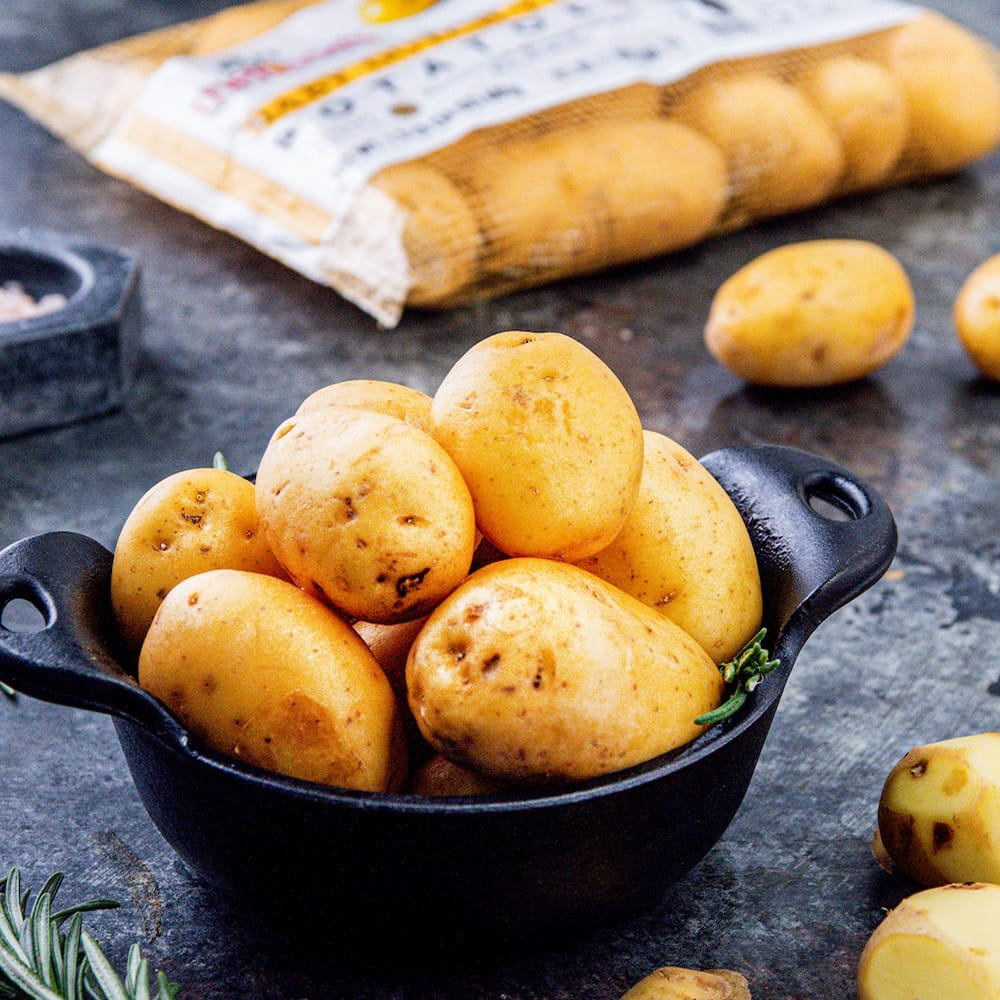 A black bowl filled with small Dutch yellow potatoes on a dark stone surface, with a bag of potatoes, loose potatoes, a peeler, rosemary, and seasoning containers arranged around it.