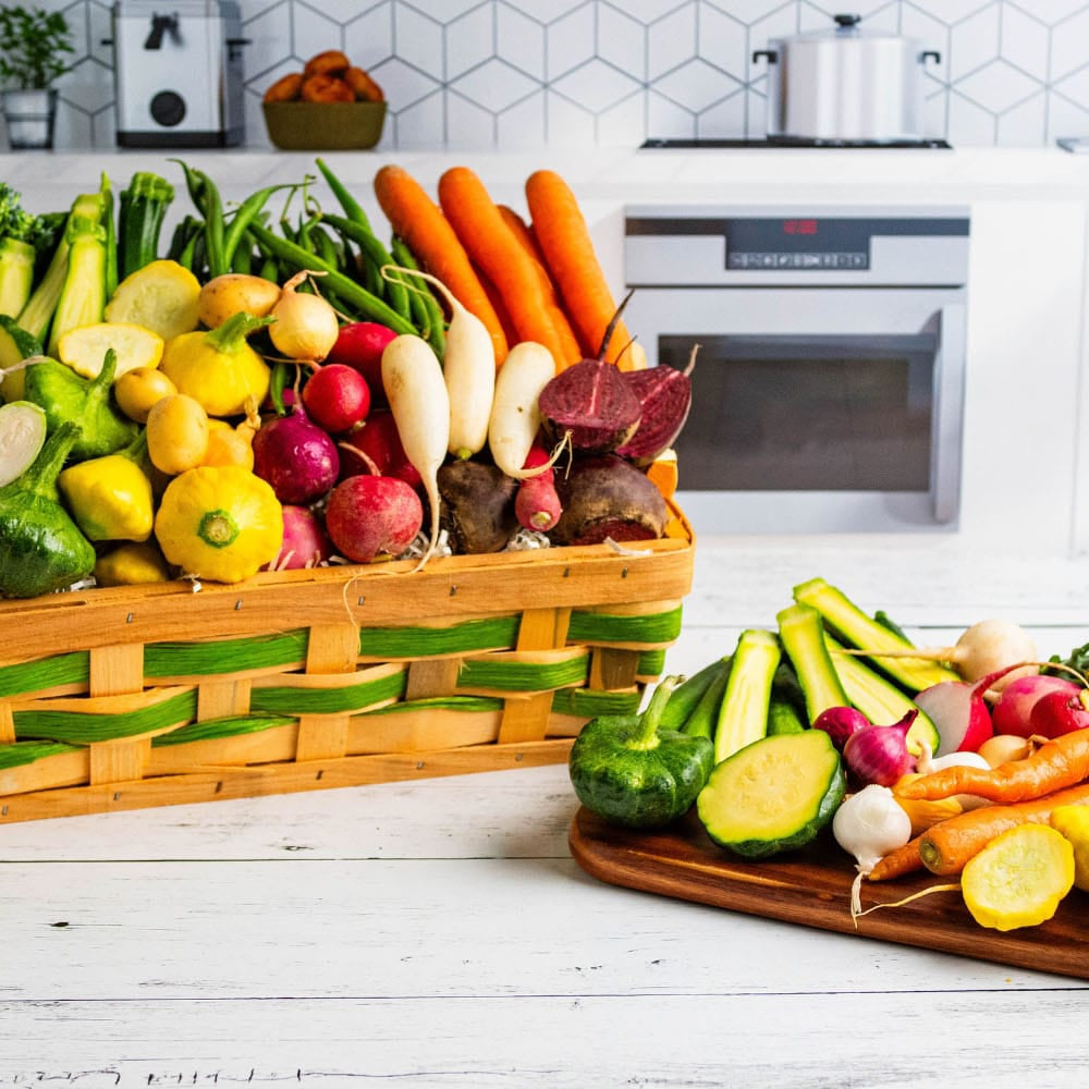 A large woven basket and a wooden cutting board filled with fresh, colorful vegetables, including broccoli, carrots, radishes, zucchini, and yellow squash, arranged on a white wooden table in a bright kitchen setting.
