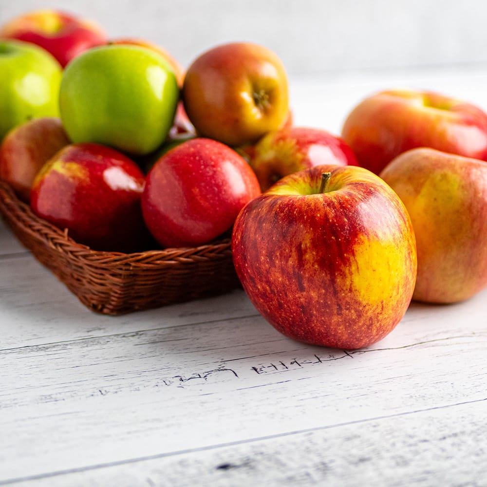A group of fresh apples in red, yellow, and green, with some arranged in a small woven basket on a white wooden surface.