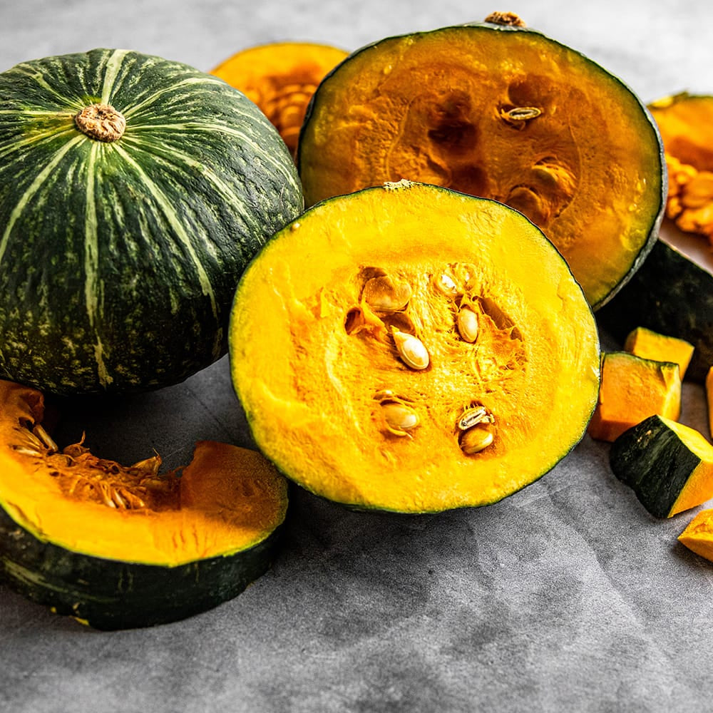 A group of kabocha pumpkins, including whole green striped pumpkins and several cut open to show the bright orange flesh and seeds, arranged on a gray stone-like surface.