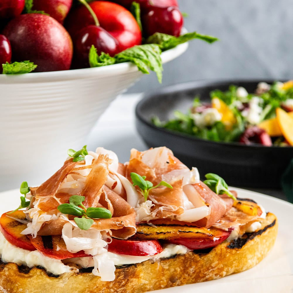 stonefruit in a white bowl, a salad with fruit and greens in a black bowl, and a plate of sliced bread topped with buratta, sliced peaches and proscuitto with microgreens.