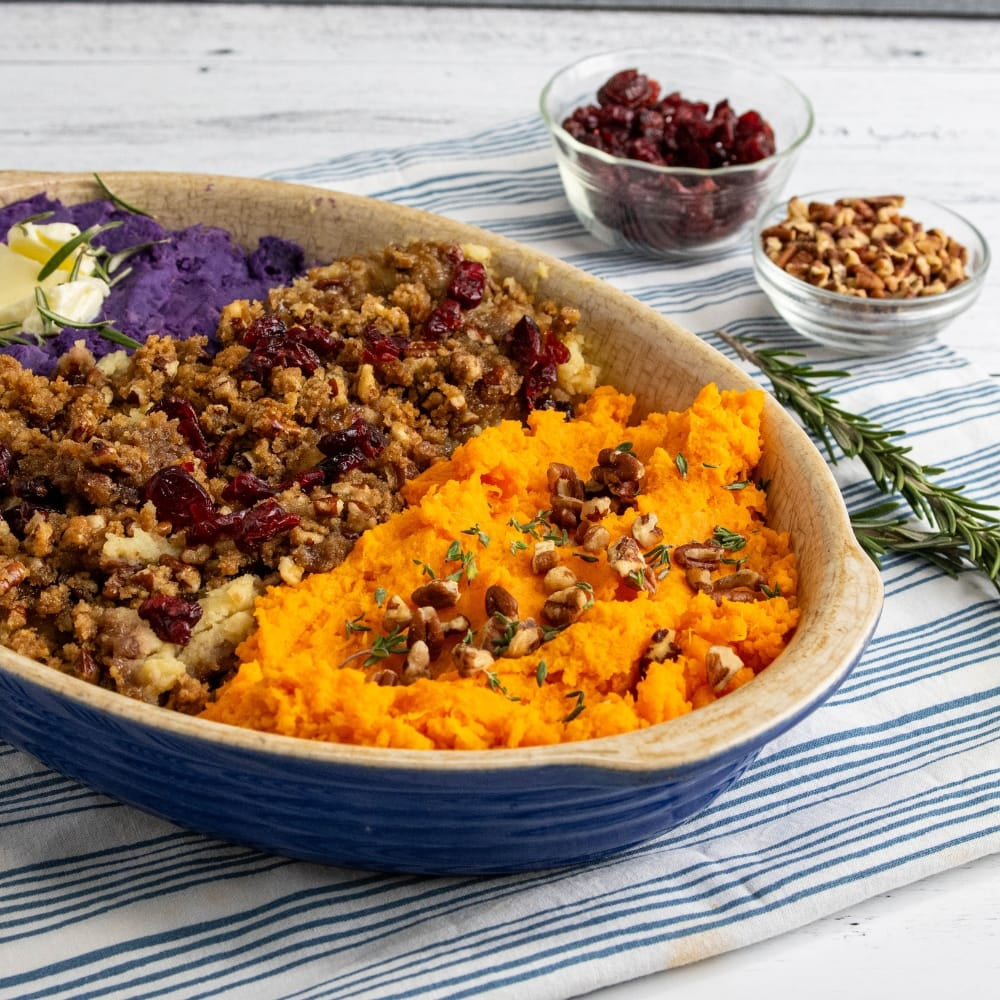 A blue oval ceramic baking dish filled with three distinct sweet potato or squash mash sections: purple, browned with cranberries and pecans, and orange. The dish is set on a blue and white striped napkin on a white wooden table, with garnishes including a sprig of rosemary, dried cranberries, and pecans.