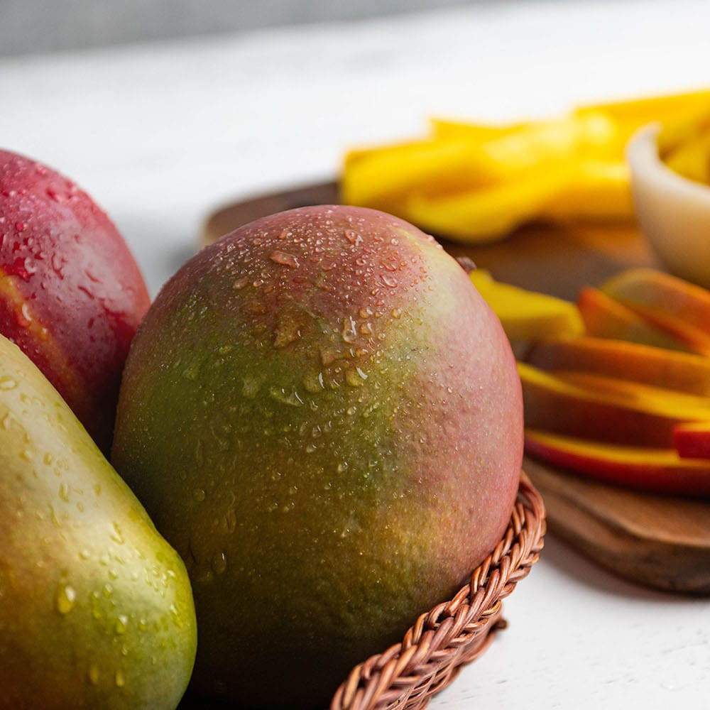 Mangoes whole and sliced on a cutting board and chunks in a bowl on a white surface