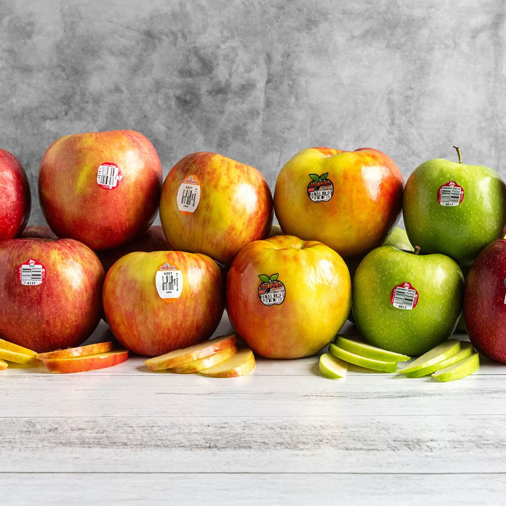 A group of fresh Gala apples in red, yellow, and green, arranged on a white wooden surface with sliced apples in front, against a textured gray background.